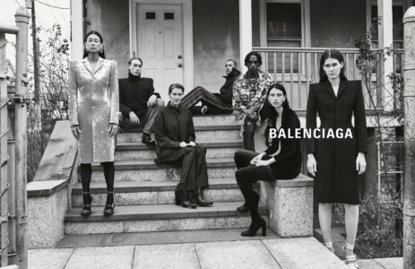 A group of seven individuals poses on the steps of a porch. Each person showcases distinct, stylish attire ranging from elegant coats to patterned outfits. The setting appears residential, with a prominent door and railings framing the scene. The composition conveys a sophisticated, contemporary vibe through both the fashion and the serious, poised expressions of the individuals.