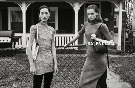 Two individuals stand in front of a house with a decorative porch. They wear stylish, textured outfits, one in a knee-length dress and the other in a longer garment. Both carry handbags. A chain-link fence separates them from the foreground. The scene captures a serene yet sophisticated ambiance, highlighting the fashion against a quaint residential backdrop.