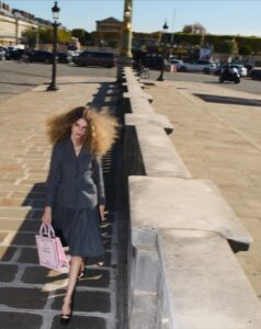 A person with voluminous hair wears a tailored grey outfit while walking along a sunlit, paved street, carrying a pink bag. The setting appears urban, featuring classic-style lampposts, vehicles, and bicycles. The background has an elegant, historical building amidst greenery, under a clear blue sky, suggesting a cobblestone walkway in a bustling city environment.