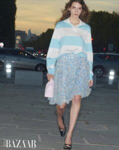A woman walks confidently on a paved urban street, dressed in a blue and white striped pullover paired with a shimmering blue skirt. She holds a light pink bag, while her feet are adorned with black heels. The background features moving cars, illuminated streetlights, and lush green trees under a dusky, softly glowing sky.