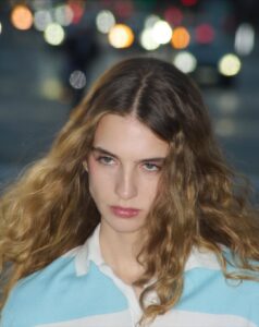A person with long, wavy, light brown hair is in focus, wearing a blue and white striped shirt. The background is out of focus, showing colorful, bright circular lights, creating a bokeh effect, suggesting a nighttime urban setting. The expression is neutral, and the composition highlights the contrast between the subject and the vibrant backdrop.