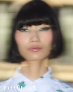 A person with a sleek black bob haircut and bangs gazes ahead. Bright green eye makeup contrasts with their subtle pink lip gloss. The background blends with a soft, dreamy bokeh effect, creating an ethereal atmosphere. Their clothing features a light pattern with hints of green, adding to the fresh, modern aesthetic.