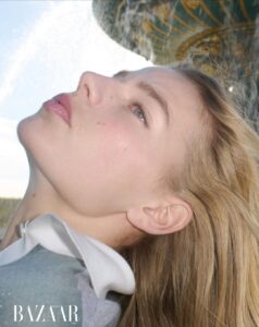 A person is portrayed in close-up from a side angle with their head tilted back, showcasing delicate features and light hair cascading around the shoulders. A soft, blurred background hints at an abstract structure with curved patterns. The serene, upward gaze suggests contemplation, with bright lighting emphasizing smooth skin and natural beauty.