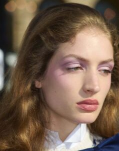 A person with long, wavy brown hair gazes thoughtfully to the side. Their expression is calm and serene, enhanced by soft pink makeup on the eyelids and a hint of blush on the cheeks. The lips are subtly colored, complementing the overall look. The attire features a high-collared white shirt, exuding a classic and elegant style.
