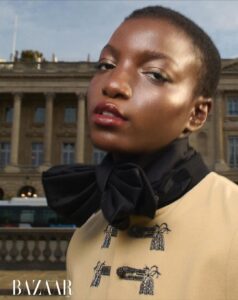 A person with short hair gazes confidently. Their makeup enhances their features with a glossy finish. They wear a beige outfit with intricate black embellishments and a large black bow around the neck. The background reveals an old building with classical architecture, including columns and arches, under a clear sky, adding an elegant urban atmosphere.