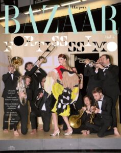 A model stands confidently at center, wearing a voluminous, sculptural dress with bold blocks of color and exaggerated shape. Around her, a group of musicians in formal black suits play brass instruments, some kneeling, others standing, creating a sense of movement and sound. The indoor setting feels theatrical, with warm lighting and a playful contrast between high fashion and lively performance.