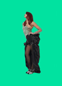 A person with long red hair stands confidently, wearing a strapless, light green top paired with a voluminous black skirt. The skirt is artfully draped and features a high slit, revealing sheer black stockings and black-and-white shoes. The pose is elegant, with one arm resting on the waist, set against a vibrant, contrasting light green background.