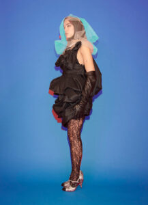 A person stands against a vibrant blue background, wearing an avant-garde outfit. The ensemble includes a voluminous black dress with ruffled layers and exaggerated sleeves. The look is complemented by elbow-length black gloves, sheer lace tights, and silver high-heeled shoes. A netted, multicolored headpiece adds a playful, artistic touch to the overall ensemble, blending fashion and art.