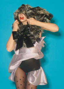 A person poses against a bright blue background wearing an avant-garde outfit. The ensemble includes layers of fabric, a large black bow, and textured elements resembling fur. Their head is partially obscured by the voluminous clothing, and they have red lips drawn on their face. Sheer polka-dotted stockings add to the creatively bold and dramatic style.