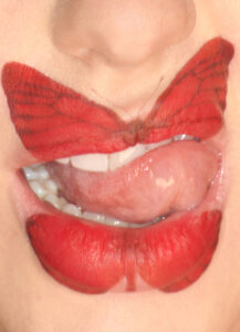Bold red lipstick forms the shape of a butterfly across lips, with intricate wing-like patterns. A tongue partially sticks out, adding a playful and unconventional touch. The upper and lower lips are colored to form the butterfly's wings, showcasing a creative and artistic approach to makeup. Teeth are slightly visible, enhancing the overall dynamic composition.