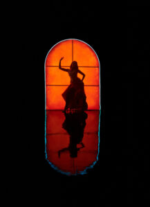 A silhouette of a person striking a dynamic pose stands against a vibrant, glowing orange backdrop, shaped like an arched window. The reflection below amplifies the silhouette, creating a symmetrical effect. The scene conveys a sense of mystery and artistry, focusing on the interplay of light and shadow in a minimalistic dark setting.