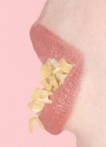 A close-up view focuses on parted lips, with a shade of soft pink, slightly glossy in texture. Nestled between them are small, randomly shaped bits of macaroni, their pale yellow contrasting gently against the lips. The background is a uniform pastel pink, enhancing the subtle, artistic quality of the composition.