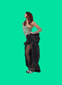 A person strikes a dramatic pose against a vibrant green backdrop, gazing upwards. They wear a strapless top with an intricate design paired with a voluminous black skirt featuring ruffled details. Her legs are visible through black stockings, and she wears white, embellished shoes. Long, flowing red hair completes her striking and fashionable appearance.