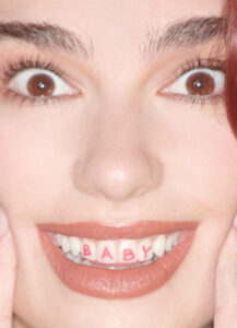 A person showcases a playful expression with wide eyes and a broad smile. Each tooth bears a pink letter, collectively forming the word "BABY." The complexion is light, and the lips are distinctly lined, enhancing the smile's vibrancy. The person's eyebrows are full, adding to the expressive appearance, and the overall tone is whimsical and lively.