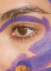 A close-up of a person's eye showcases intricate details of the eyelashes, eyebrow, and iris. The surrounding skin is adorned with bold, vibrant strokes of purple paint, creating a striking contrast. The purple hues sweep across the brow and cheekbone, highlighting the eye's deep, contemplative gaze and adding an artistic flair to the overall composition.
