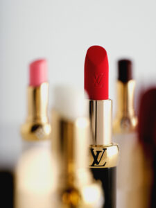 A collection of elegant lipsticks in various vibrant shades stands on a soft, blurred background. The main focus is a red lipstick with gold accents, surrounded by others in different hues such as pink and deep burgundy. The lighting reflects subtly off their shiny surfaces, emphasizing their luxurious and sophisticated appeal.
