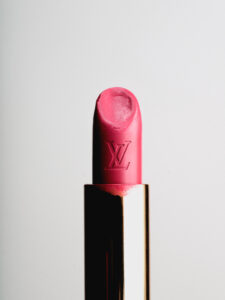 A vibrant pink lipstick is elegantly displayed with a smooth, round top and subtle markings on the surface. Its golden casing provides a luxurious contrast, enhancing the rich color. The lipstick shows slight use, indicated by a small imprint. The backdrop is a plain, soft white, allowing the boldness of the lipstick to stand out prominently.