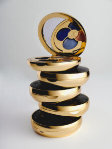 A stack of round, shiny gold compacts is neatly arranged. The top compact is open, revealing a mirror and an eyeshadow palette with deep blue and warm brown shades. The sleek, reflective surfaces create an impression of luxury and elegance, while the overall composition conveys a sense of sophistication and high-end beauty products.