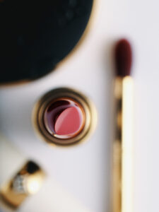 A luxurious arrangement showcases makeup essentials with a focus on a vibrant pink lipstick, captured from above. The lipstick is surrounded by a golden container, while a soft-focus brush lies adjacent, featuring a matching hue. The background is a clean, white surface, enhancing the elegance and sophistication of the cosmetic items.