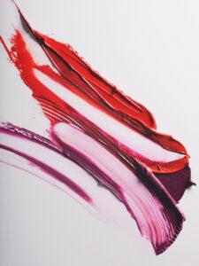 Vivid smears of color sweep dynamically across a clean surface, blending hues of red, pink, and purple. The thick texture and bold shades contrast against the light background, creating a striking visual. The overlapping strokes suggest motion and energy, as vibrant reds mix harmoniously with softer purples, generating a sense of artistic fluidity and expressive creativity.