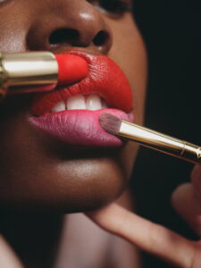 Close-up of a person applying makeup to lips. The lips are painted with bright red and pink colors. A red lipstick is being applied to the upper lip, while a brush touches the lower lip. The skin tone is deep and smooth, with a soft focus on the glowing texture, emphasizing the vibrancy of the makeup.