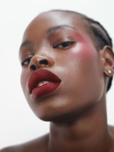 A close-up, artistic portrait showcases a person with smooth skin and a poised expression. The individual wears bold, red lipstick and subtle eye makeup that highlights their features. Soft lighting emphasizes the sheen on their skin, adding depth and texture. Their hair is styled neatly, complementing the overall polished and elegant appearance. A gold earring adds a touch of sophistication.