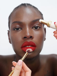 A person with deep red lipstick has makeup being applied. A makeup brush touches the lip area, adding a lighter tone, while another tool is near the eyebrow. The background is neutral, enhancing focus on the face. The person’s skin is smooth, and the overall tone is warm and vibrant, highlighting their expressive eyes and striking features.