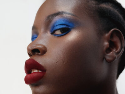 A person gazes upward with a captivating expression, featuring vibrant blue eyeshadow and bold red lipstick. The skin glows with a natural sheen, and the close-up perspective highlights facial features and textures. The hair is styled in neat, subtle braids, and the overall tone suggests a blend of elegance and drama in the presentation.
