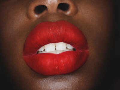 A close-up showcases vibrant red lips, slightly parted to reveal white teeth. The smooth skin surrounding the lips contrasts sharply with the bold lipstick, highlighting the rich color. The upper portion subtly includes the lower part of a nose, emphasizing the focus on the lips. The composition is striking, with deep colors creating an impactful visual.