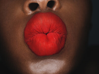 Vibrant red lips puckered as if blowing a kiss, set against a smooth, dark complexion. The focus is tight, capturing the luscious texture and shine of the lipstick. There's a sense of intimacy and boldness conveyed through the contrasting colors. The image captures a moment that feels both playful and sensual, emphasizing the lips' natural beauty.