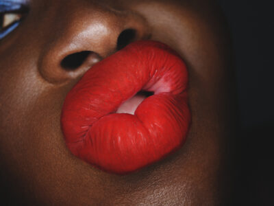 A close-up depicts a person's full, glossy red lips slightly parted, forming an expressive pout. The vibrant red lipstick contrasts vividly against the person's dark, smooth skin, creating a bold, striking effect. The detail highlights the texture and sheen of the lips, conveying a sense of glamour and intensity, enhanced by a subtle play of light and shadow.