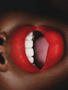 A close-up view of open lips showcases vibrant red lipstick, with a focus on the teeth and the contrasting dark background. The bright red hue is striking against the rich skin tone, emphasizing the textures and curves of the lips. The composition captures a sense of boldness and intimacy, drawing attention to the vivid and dramatic colors.