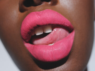 A close-up reveals a set of lips adorned in vibrant pink lipstick, slightly parted to reveal white teeth. The skin has a rich, smooth texture, highlighted in natural lighting. The overall effect is striking and bold, with the vivid color of the lipstick drawing attention and adding a touch of glamour and sophistication.