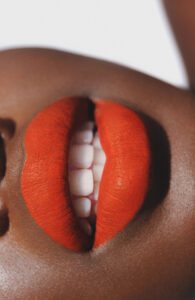 Lush red lips prominently feature in the center, with a hint of white teeth showing beneath. The rich, smooth texture of the lips contrasts against the warm brown skin, creating a captivating focal point. The close-up perspective emphasizes the vibrant color and fine details, offering an intimate, striking representation of beauty and self-expression.