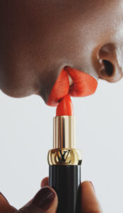 Bold red lips are being carefully colored with a vibrant red lipstick. A hand with neatly manicured nails holds the lipstick, accentuated by a gold band with engraved letters. The composition emphasizes the rich contrast between the deep skin tone and the vivid lip color, creating a striking and elegant visual effect.
