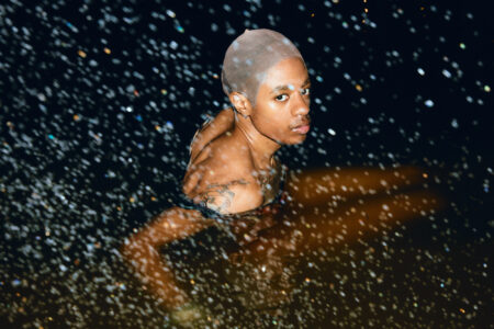 A person is partially submerged in water, looking directly at the viewer. The dark surroundings contrast with the scattered reflections and droplets. The individual has tattoos on their shoulder and wears a sheer head covering. The lighting creates a dramatic effect, highlighting the person's features and the dynamic play of light on the water surface.