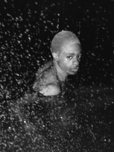 A person with a shaved head is submerged in dark water, illuminated by overhead lighting. Their gaze is direct and intense, creating an evocative contrast with the surrounding darkness. Water droplets are visible in the foreground, adding texture to the scene. The overall mood is mysterious and contemplative, enhanced by the interplay of light and shadow.