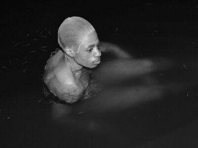 A person with a shaved head is submerged in dark water, visible from the shoulders up. Their focused expression is captured in profile, with light and shadows accentuating their facial features. Small, indistinct objects appear to float around them. The monochromatic setting emphasizes the calm and introspective mood of the scene.