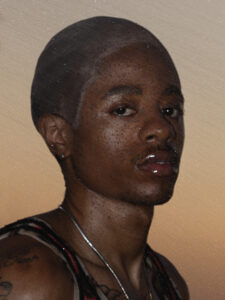 A person gazes directly ahead, with a serious expression. The skin glistens with tiny water droplets, creating a rain-drenched effect. An orange and brown background suggests a warm, subdued lighting, possibly during sunset. They wear a sleeveless garment and a necklace, adding a stylish element along with visible tattoos on the arm, enhancing personal expression.