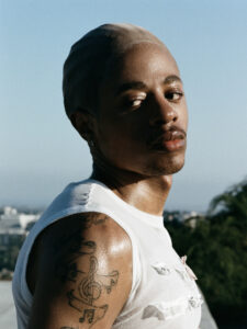 A person gazes into the distance, wearing a fitted cap and sleeveless white top. Sunlight highlights their features and tattoos featuring musical symbols on the arm. The background shows a blurred cityscape and greenery under a clear blue sky. The overall mood conveys a sense of calm and introspection amid the urban environment.