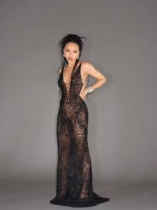 A person poses confidently, wearing a long, intricately designed sheer black dress with a plunging neckline. The garment features a delicate web-like pattern. Their hair is styled upwards with extensions, and they accessorize with silver bracelets. The background is simple and gray, emphasizing the stylish and avant-garde outfit and relaxed, poised demeanor.