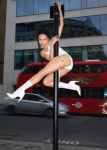 A person is striking an acrobatic pose on a pole along a city street. Dressed in stylish attire with high-heeled boots, they exude confidence. Behind them, a red double-decker bus and a sleek car are in motion. The backdrop features a building with reflective windows, capturing urban life in a lively, dynamic scene.