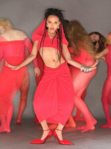 A person stands confidently, wearing an artistic red outfit with matching heels. Their hair is styled in braids, and they accessorize with large hoop earrings and multiple bracelets. Behind them, several individuals dressed in red bodysuits move dynamically, creating a sense of energy and motion. The scene conveys a vibrant and creative atmosphere, emphasizing bold fashion.
