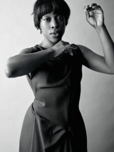 A person with short hair poses gracefully, captured in a monochrome setting. They wear a sleeveless dress featuring a subtle knot detail at the waist. One arm is bent near the face, while the other is raised, conveying a sense of movement and elegance. The lighting highlights the textures and contours, creating a striking visual composition.