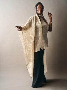 A person stands confidently against a neutral background, wearing a flowing, striped, light-colored garment with wide sleeves. Their attire extends downwards, contrasting with darker pants. The individual has an elegant posture with one arm outstretched, and their hair is styled with braids or twists. They wear minimal jewelry, including a wristwatch, enhancing their graceful and poised appearance.