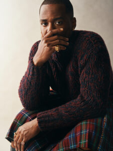A contemplative individual sits, their hand partially covering their mouth. They wear a textured, dark sweater with intricate detailing and a gold ring with a lion motif. A plaid fabric in rich colors drapes over their lap. The person’s gaze is focused and intense, set against a softly lit, neutral background, creating a serene yet striking atmosphere.