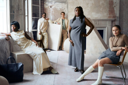 Five individuals pose with confidence in an elegantly styled, rustic interior. Their outfits vary from sleek dresses to cozy knits, showcasing a blend of textures and neutral tones. The setting features distressed walls, large windows, and an ornate fireplace, creating an artistic, modern atmosphere. Stylish accessories, such as boots and a leather bag, add sophistication to the scene.