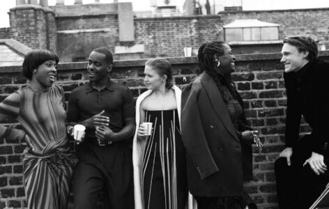 A group of five people stand together on a rooftop against a brick wall, engaged in lively conversation. One person on the left has short hair and is gesturing animatedly. Others hold cups, suggesting a casual gathering. The scene exudes a relaxed and friendly atmosphere, with everyone dressed in stylish attire, enjoying each other's company.