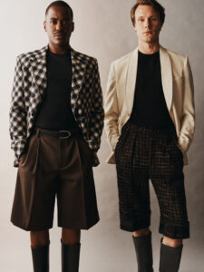 Two individuals stand confidently against a neutral background, displaying contrasting yet complementary fashion styles. On the left, a person wears a checkered blazer and dark knee-length shorts with tall boots. On the right, the person sports a cream blazer and patterned shorts, also wearing boots. Both exhibit a modern, sophisticated look with neutral-toned tops, exuding elegance and style.
