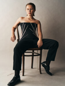 A person with short hair sits confidently on a wooden chair against a neutral backdrop, wearing a strapless top adorned with striking linear embellishments. The outfit is completed with sleek black trousers and polished shoes, projecting an air of sophisticated nonchalance. The overall mood combines elegance with a casual, self-assured presence, highlighting a blend of modern fashion aesthetic.